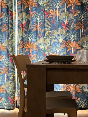 Un'area pranzo con tavolo e sedie, una ciotola sul tavolo e una tenda con motivo a foglia tropicale.