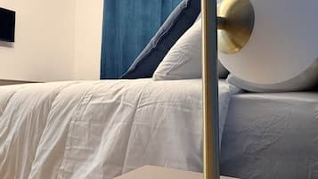 Un lit avec une literie blanche et un rideau bleu, une table ronde avec un pied doré et un téléviseur fixé au mur.