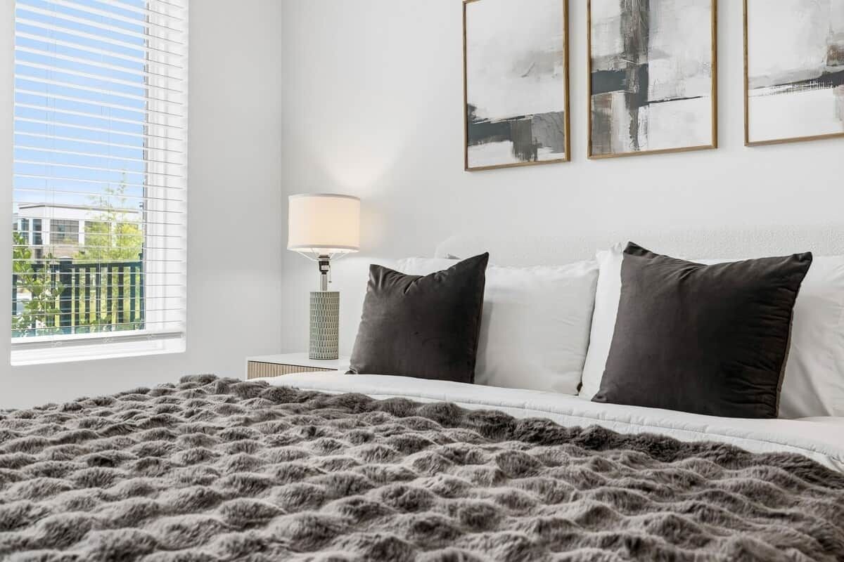 A bedroom with a large bed, two pillows, a fluffy blanket, a bedside lamp, and three abstract paintings on the wall.