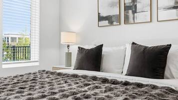 A bedroom with a large bed, two pillows, a fluffy blanket, a bedside lamp, and three abstract paintings on the wall.