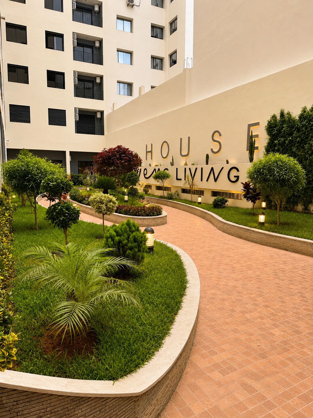 Een hotelentree met een aangelegde tuin en een bord waarop 'HOUSES LIVING' staat.