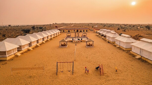 A desert resort with multiple white tents arranged in rows, a central building with a flat roof, and a playground with swings and a slide. - Experience the Best Jaisalmer Desert Safari & Luxury Camp Stay (Jaisalmer)