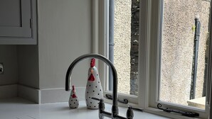 Uma cozinha moderna com bancada branca, torneira de aço inoxidável e janela com vista para uma parede de pedra.