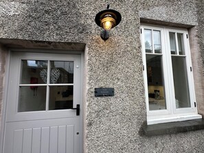 Eine Fassade aus strukturiertem Stein mit einer weißen Tür und einem Fenster, einer schwarzen Wandleuchte und einer Tafel mit der Aufschrift 'La'al Kirkstone Brow'.