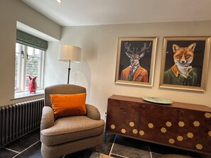 Ein Zimmer mit Fenster, einem Sessel, einer Lampe, einem Heizkörper und zwei gerahmten Bildern an der Wand.