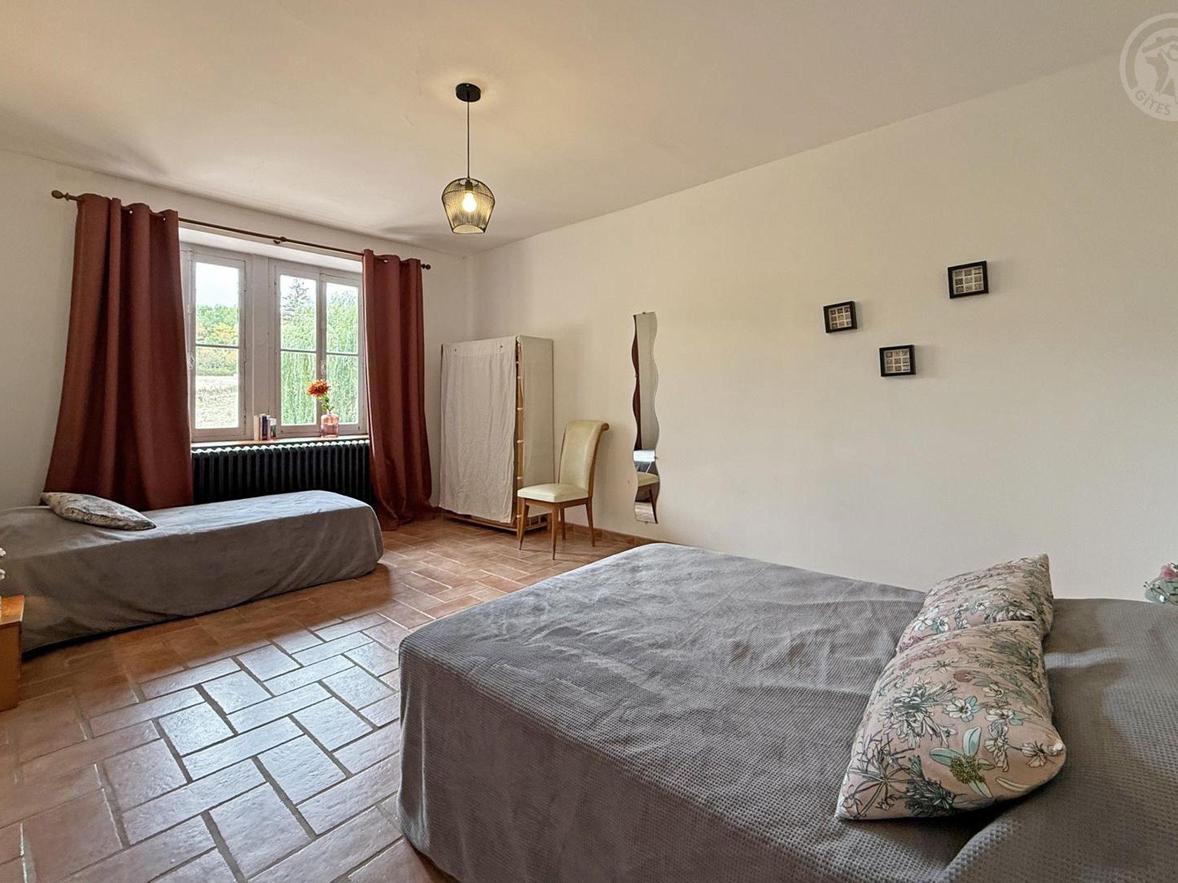 A room with two beds, a radiator, a window with curtains, a chair, and a wall-mounted mirror.