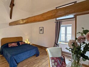 Ein Schlafzimmer mit Holzbalken an der Decke, einem Bett mit blauer Bettdecke, einem Nachttisch mit Lampe, einem Sessel mit Blumenkissen und einem Fenster mit Vorhängen.