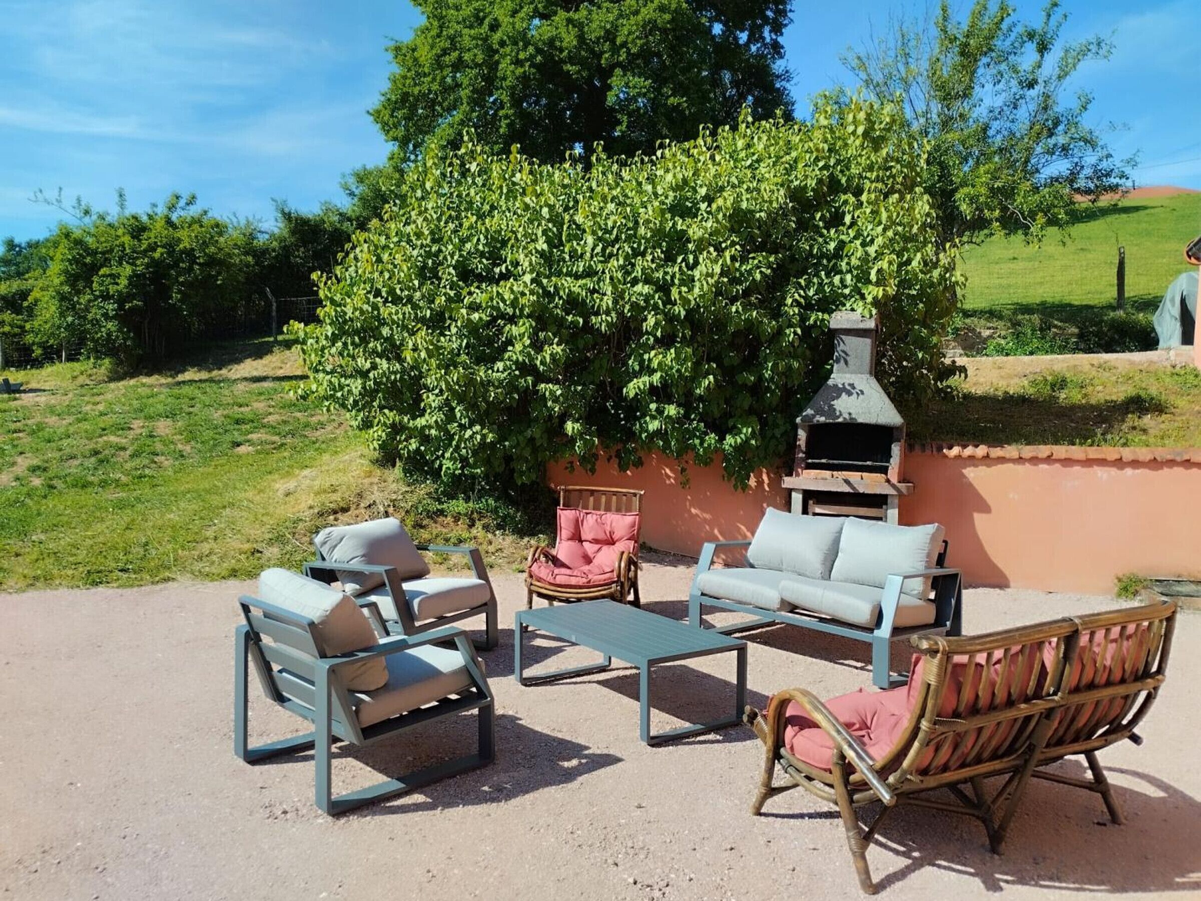 Ein Sitzbereich im Freien mit einem Grill, umgeben von viel Grün und blauem Himmel.