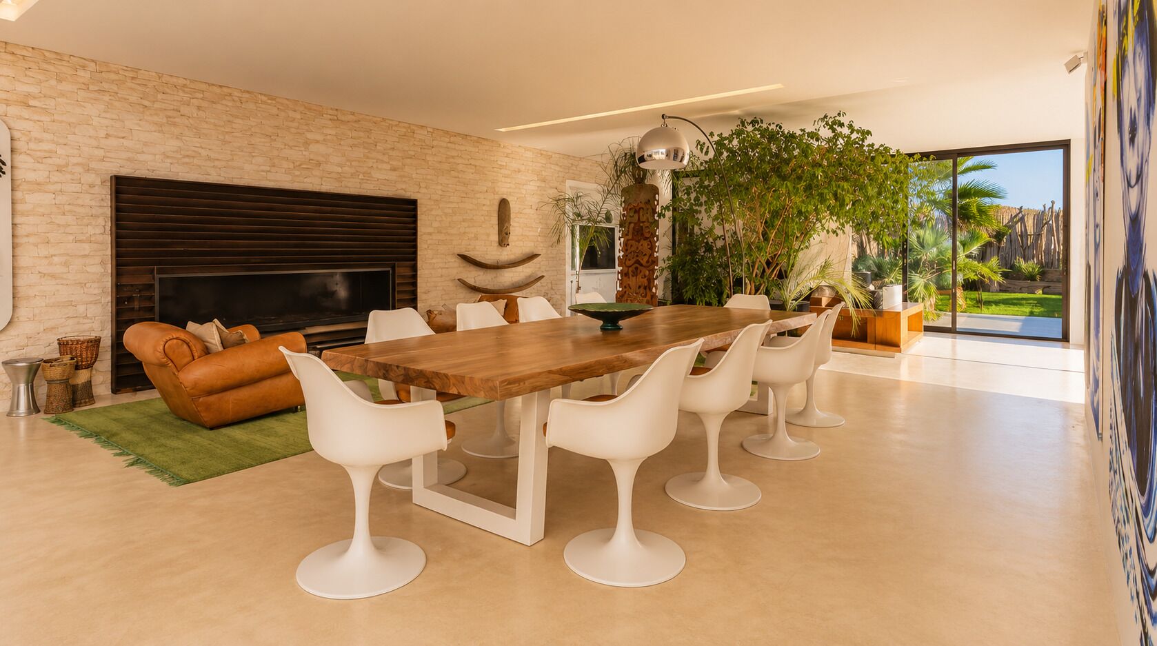 A modern living room with a fireplace, a dining table with white chairs, and a large window with a view of greenery.