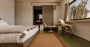 Ein Schlafzimmer mit einem Bett, einem Sessel, einem kleinen Tisch und einem Fenster mit Blick auf Kakteen.