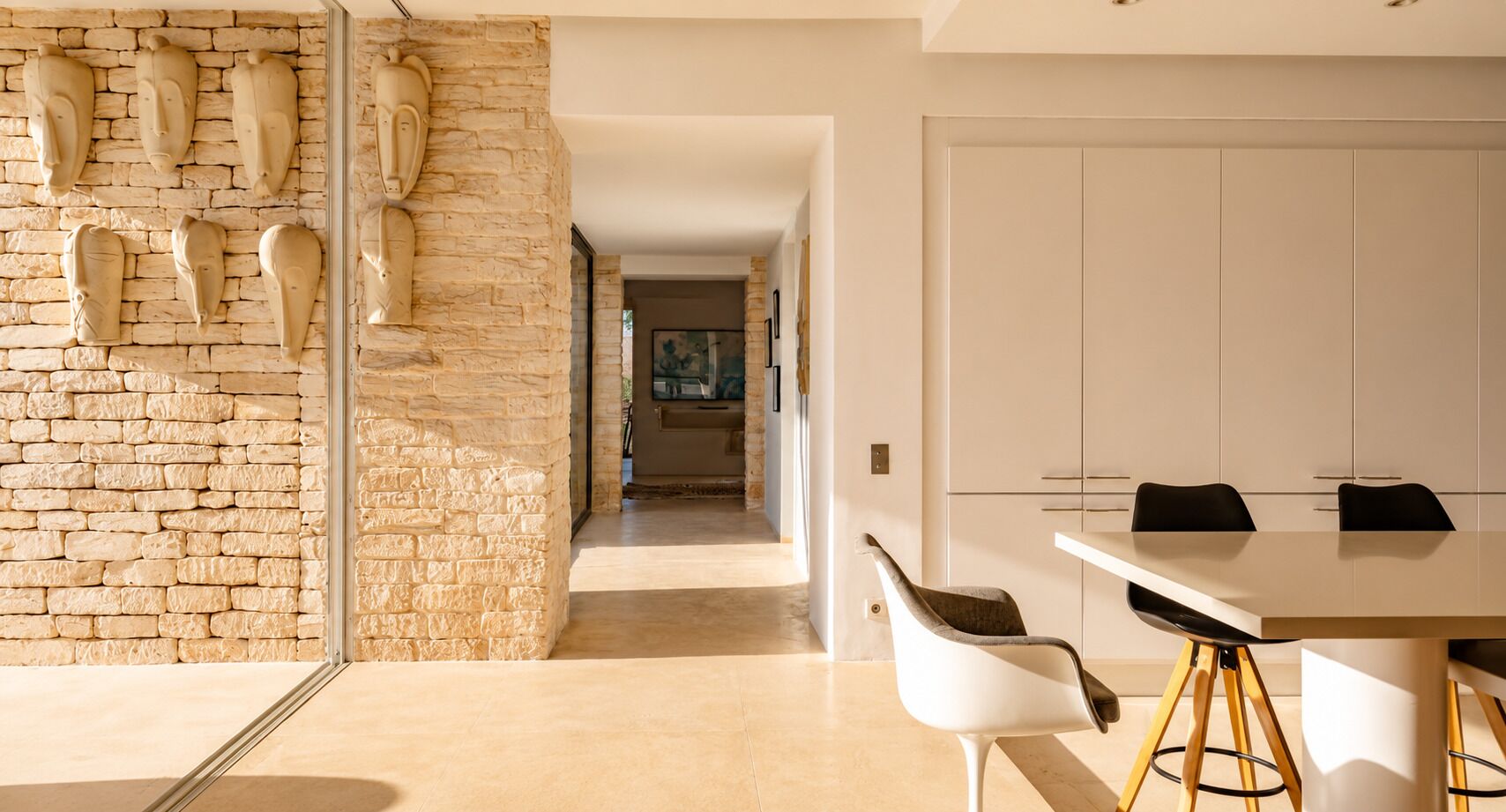 Un intérieur moderne avec un mur en pierre, un coin repas avec des chaises blanches et une cuisine en arrière-plan.