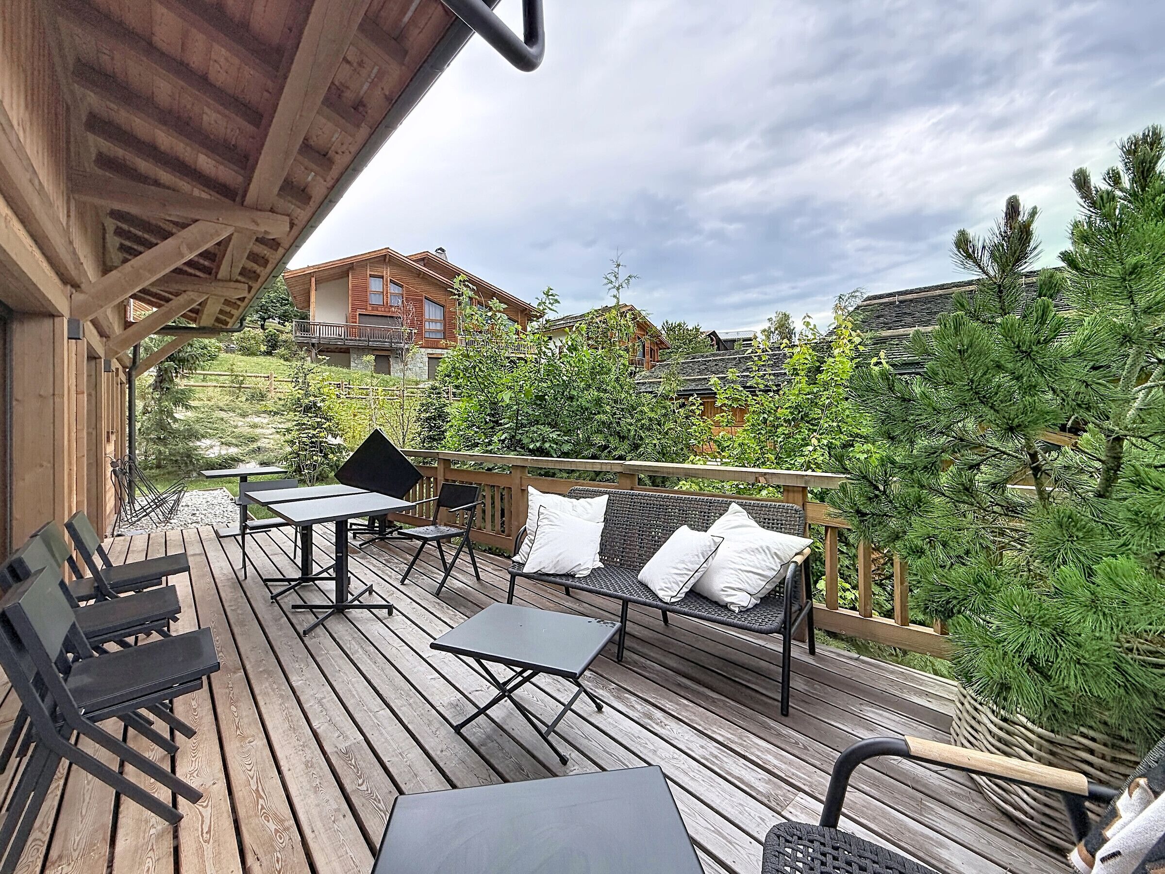 Una terraza de madera con mobiliario exterior, incluyendo mesas y sillas, da vista a un jardín con árboles y un edificio al fondo.