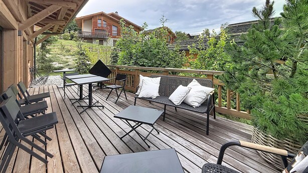 A wooden deck with outdoor seating, including tables and chairs, overlooking a garden with trees and a building in the background. - Stunning half-chalet in the village centre - 8 guests (Combloux)