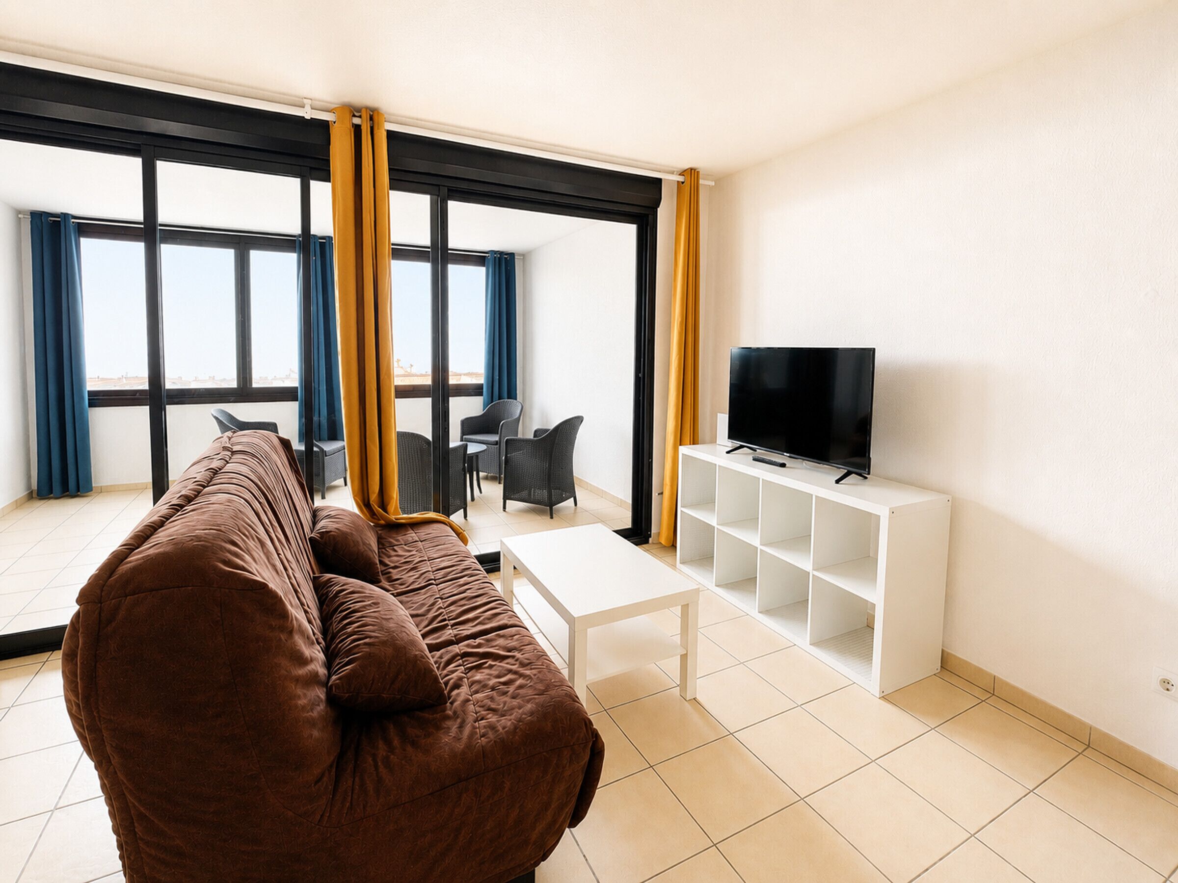 A modern living room with a sofa, a coffee table, a TV on a stand, and a balcony with a view.