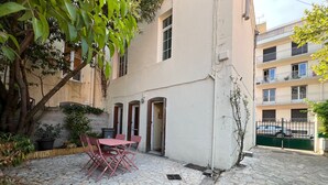 Um pátio com piso de pedra, uma mesa com cadeiras, um vaso de planta e um edifício com várias janelas e varandas.