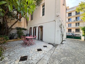 A courtyard with a stone floor, a table with chairs, a potted plant, and a building with multiple windows and balconies.