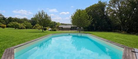 Una piscina rectangular con muelle de madera, rodeada de césped, árboles y un edificio al fondo.