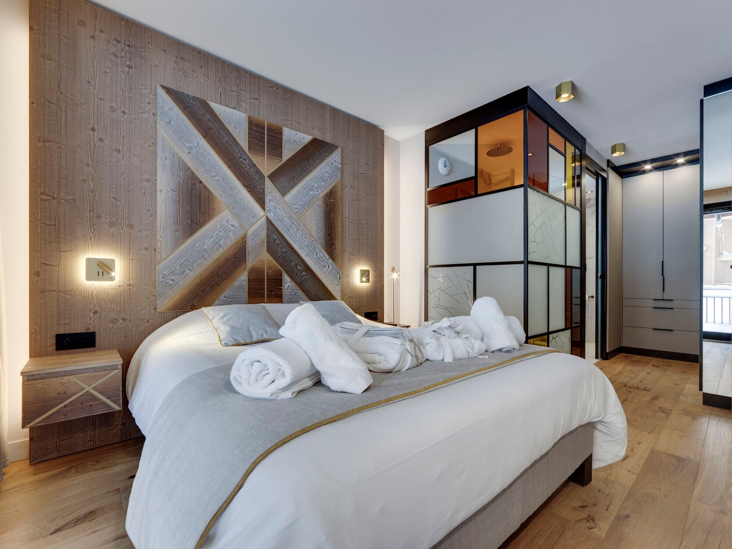 Quarto moderno com cama grande, painéis de parede de madeira e divisória de vidro.