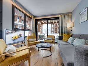 A modern living room with a sofa, armchairs, a coffee table, and a television. There are large windows with curtains, and a view of a building with balconies. - Modern apt with Spa and parking, Arc 1800 (Les Arcs 1800)