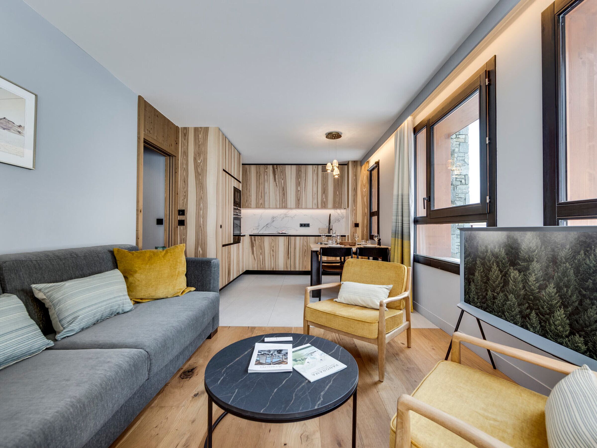 Een moderne woonkamer met een bank, fauteuil en salontafel, een eethoek met stoelen en een tafel, en een keuken met houten kasten.
