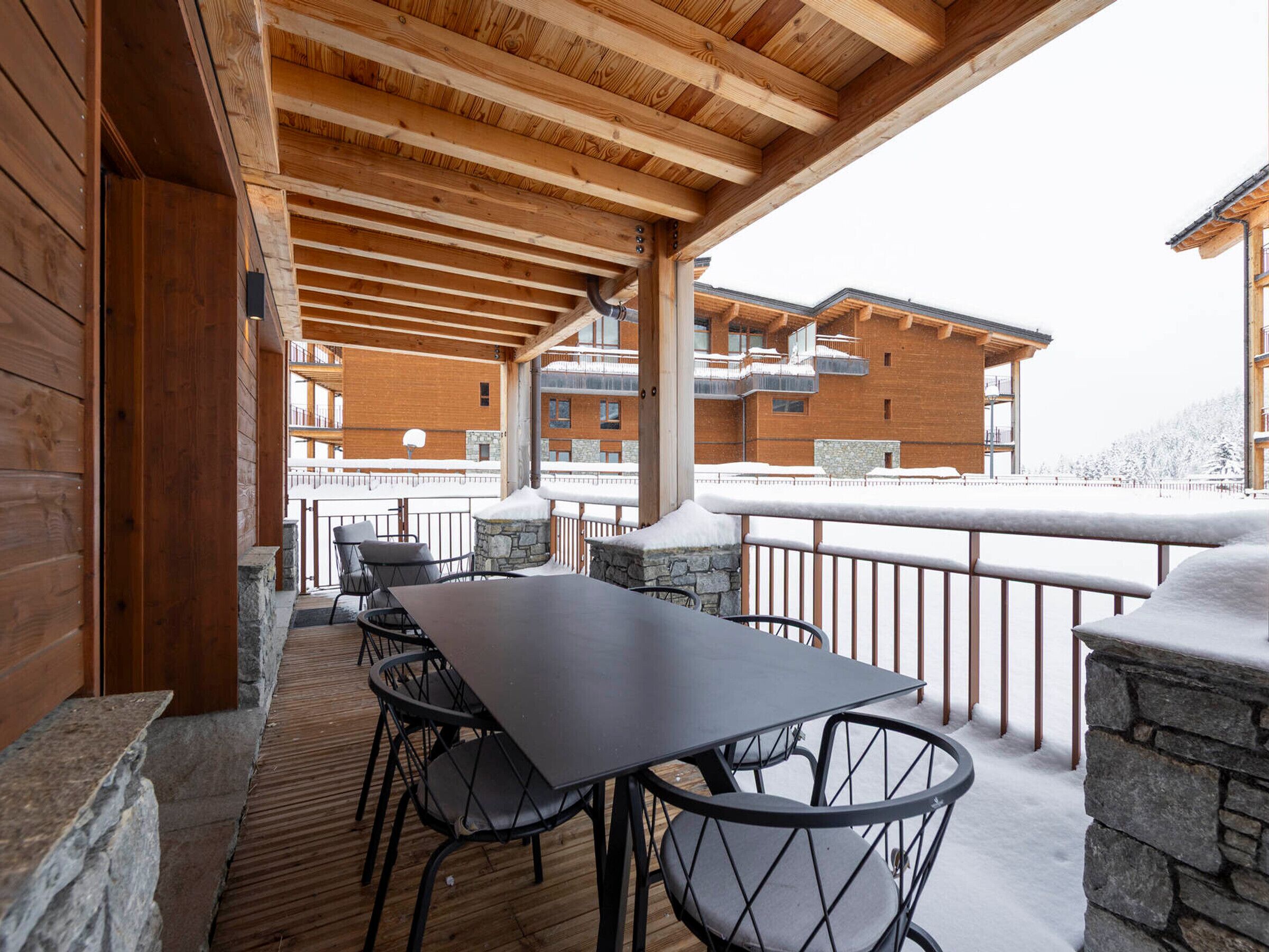 A covered outdoor seating area with a wooden ceiling, stone walls, and a view of a building with balconies.