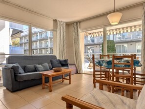 Ein Wohnzimmer mit einer Couch, einem Holztisch und einem Sessel. Große Fenster mit Vorhängen und ein Balkon mit Blick auf andere Gebäude.