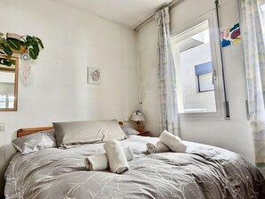 Un dormitorio con una cama, un ventanal, una planta y un cuadro colgado en la pared.