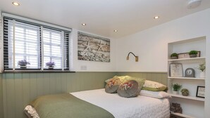A bedroom with a bed, a window with blinds, a wall-mounted shelf with decorative items, and a wall art piece. - No 1 Old Cottages (Ripon)