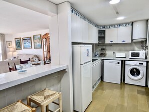 A compact kitchen with white cabinets, a refrigerator, microwave, and a washing machine. There is a dining area with a table and chairs, and a small stool. - Seafront apartment, pets - 6 guests (Tossa de Mar)