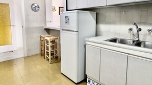A compact kitchen with white cabinets, a built-in sink, and a refrigerator. There are two stools against the wall and a door with a frosted glass panel.
