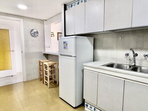 A compact kitchen with white cabinets, a built-in sink, and a refrigerator. There are two stools against the wall and a door with a frosted glass panel. - Seafront apartment, pets - 6 guests (Tossa de Mar)