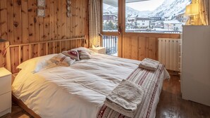 A wooden cabin bedroom with a bed, bedside tables, a window with a view of snow-covered mountains, and a radiator. - Family apartment in center, mountain view - 4 guests (Val-d'Isère)