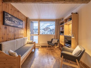Una habitación acogedora con piso de madera, un sofá, una silla, una estantería y un ventanal con vistas a una montaña nevada.