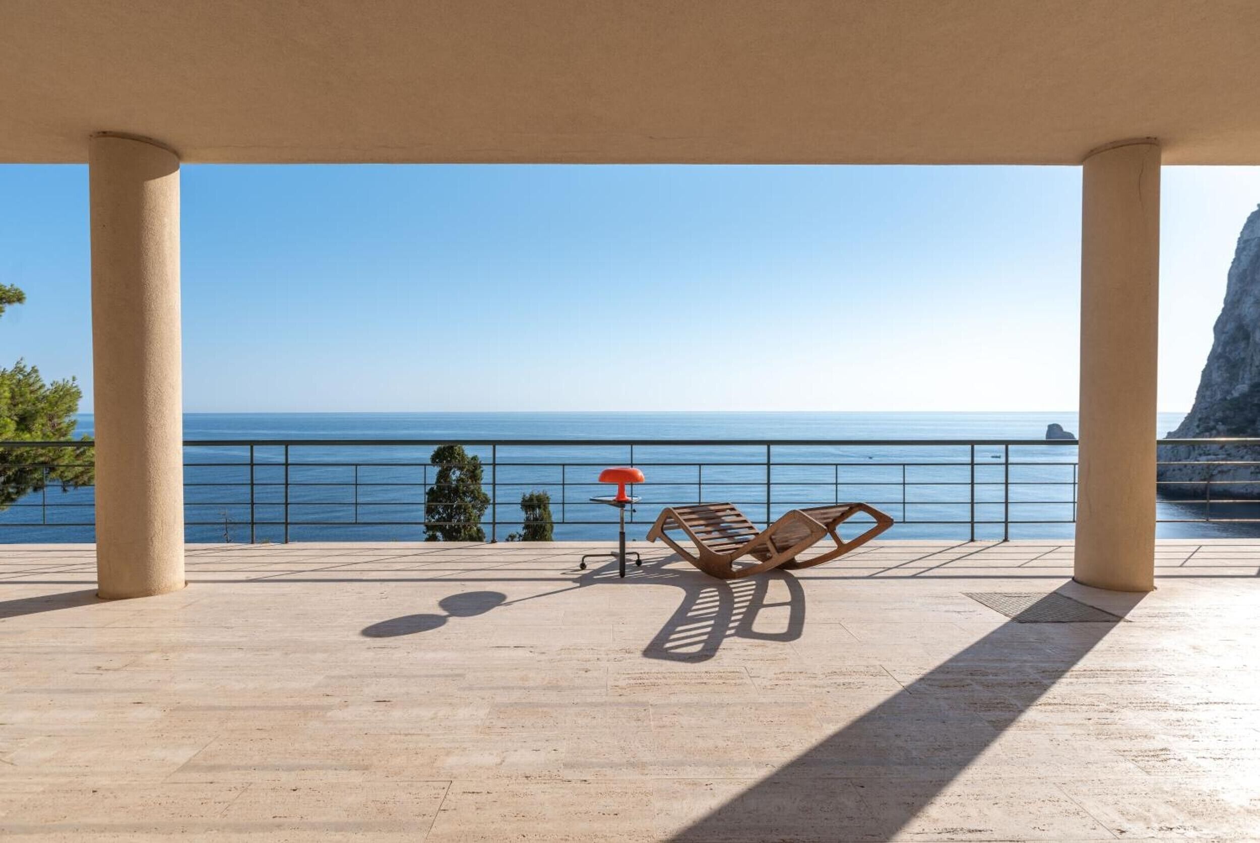 A covered outdoor area with a bench and a table, overlooking a body of water and a rocky coastline.