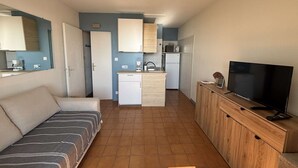 Un ambiente compatto con angolo cottura, divano, televisione e pensile in legno.