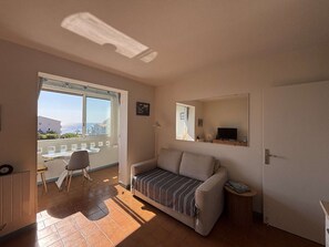 Ein Wohnzimmer mit einer Couch, einem kleinen Tisch, einem Sessel, einem Fernseher und einem Fenster mit Blick auf Gebäude.