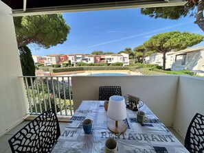 A balcony with a table set for two, a pool in the background, and a view of residential buildings. - Apartment with pool, parking - 6 guests (Cap d'Agde)