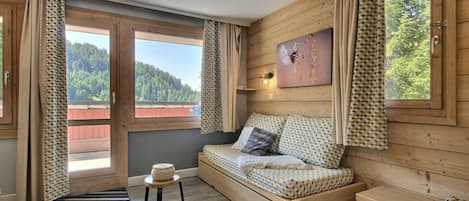 A cozy room with a wooden bed, a small stool, and a view of a forested area through large windows.