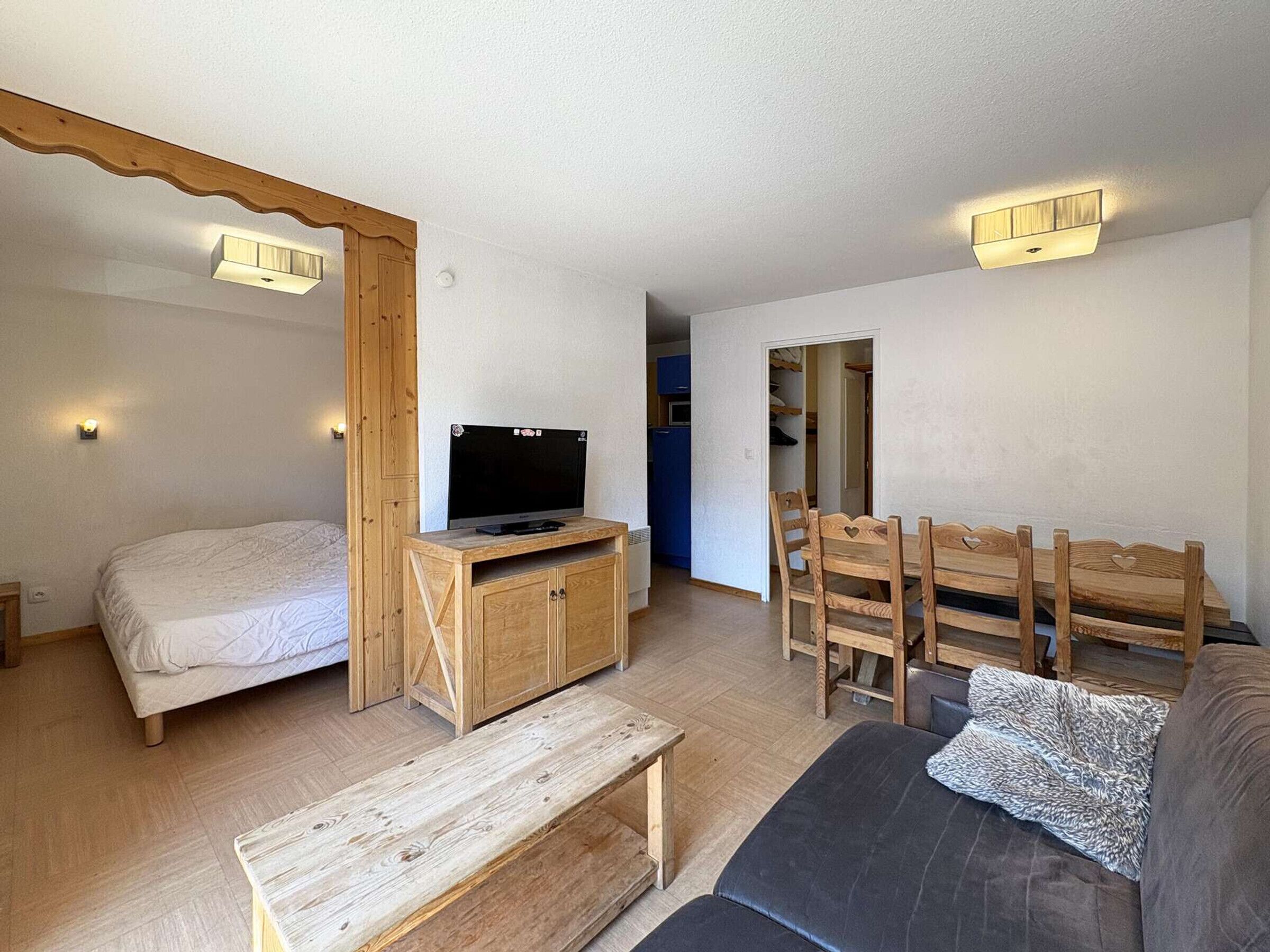 A compact living space with a bed, a television on a wooden stand, a dining table with chairs, and a sofa.