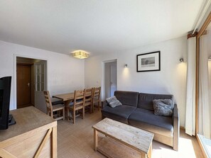 A modern living room with a sofa, coffee table, dining table, and chairs. There is a framed picture on the wall and a ceiling light fixture. - Ski-in/ski-out apartment - 8 guests (Les Orres)
