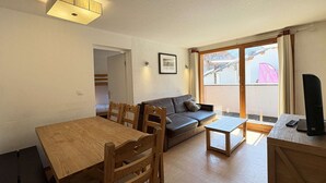 Ein Wohnzimmer mit einem Holzesstisch, einer Couch und einem kleinen Couchtisch. Ein Fenster bietet Blick auf ein Gebäude.