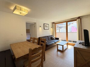 A living room with a wooden dining table, sofa, and a small coffee table. There is a window with a view of a building outside. - Ski-in/ski-out apartment - 8 guests (Les Orres)
