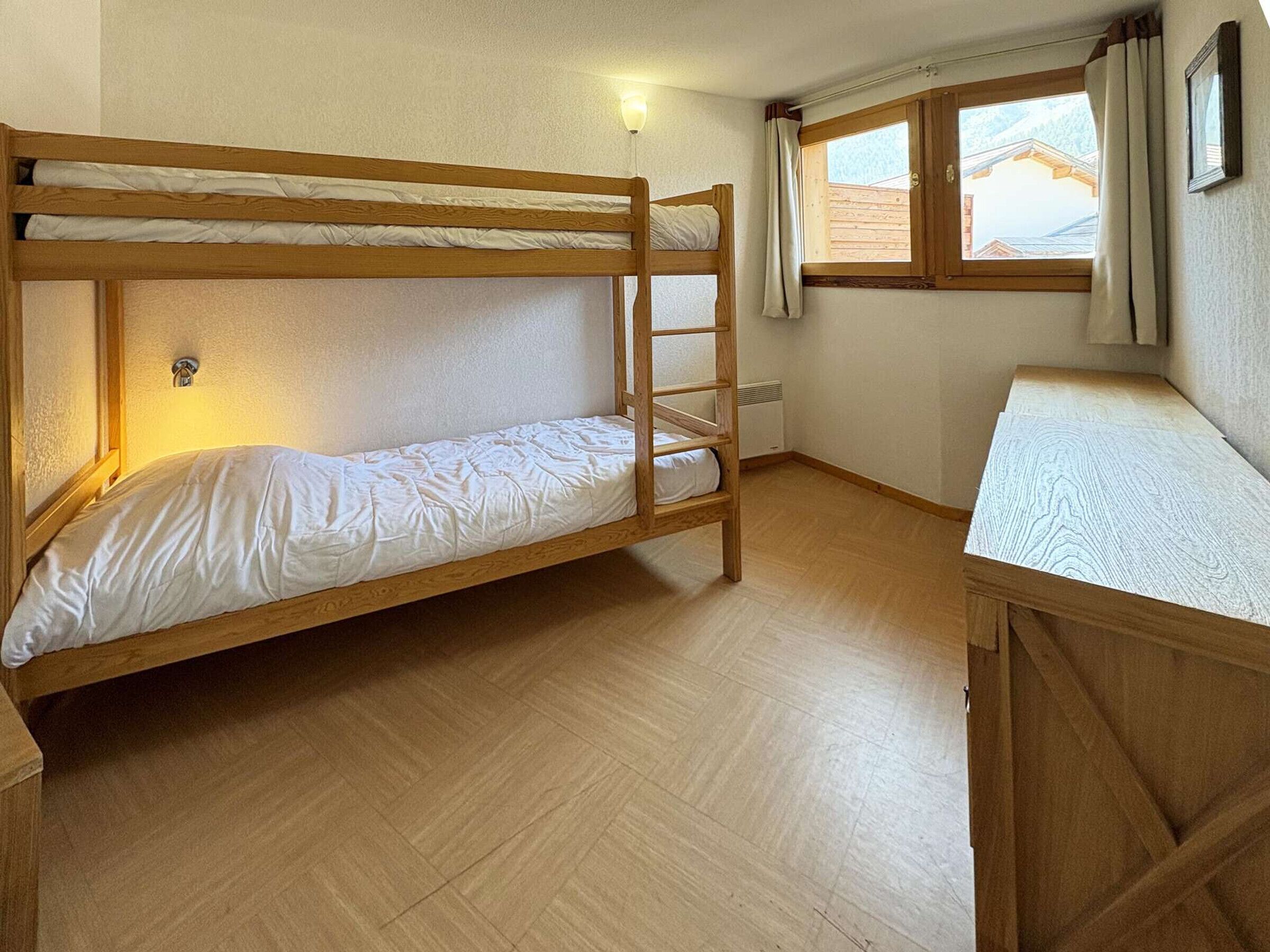 A bunk bed room with a wooden floor, a window with curtains, and a wall-mounted light fixture.