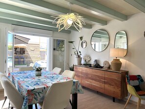 A dining area with a table covered in a floral tablecloth, surrounded by chairs, a wooden cabinet, and a hanging light fixture. - Quiet house with pool - 6 guests (Saint-Hilaire-de-Riez)