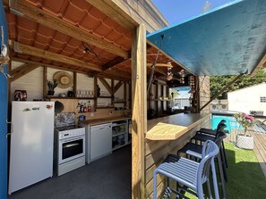 A covered outdoor kitchen with a wooden structure, a refrigerator, a stove, and bar stools. - Quiet house with pool - 6 guests (Saint-Hilaire-de-Riez)