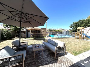 Eine Außenpatio mit Holzdeck, einer gepolsterten Couch, einem Tisch mit einem Krug und einem großen Sonnenschirm.