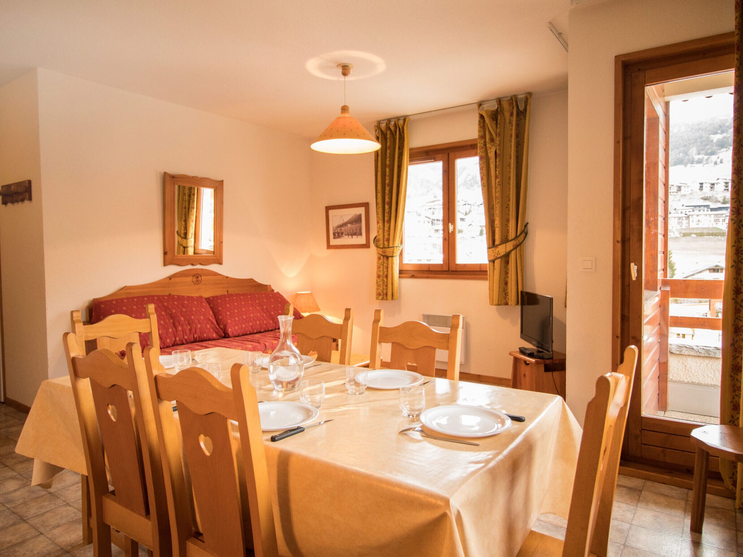 Un comedor con una mesa preparada para dos, una cama con ropa de cama roja, un televisor y una ventana con cortinas.