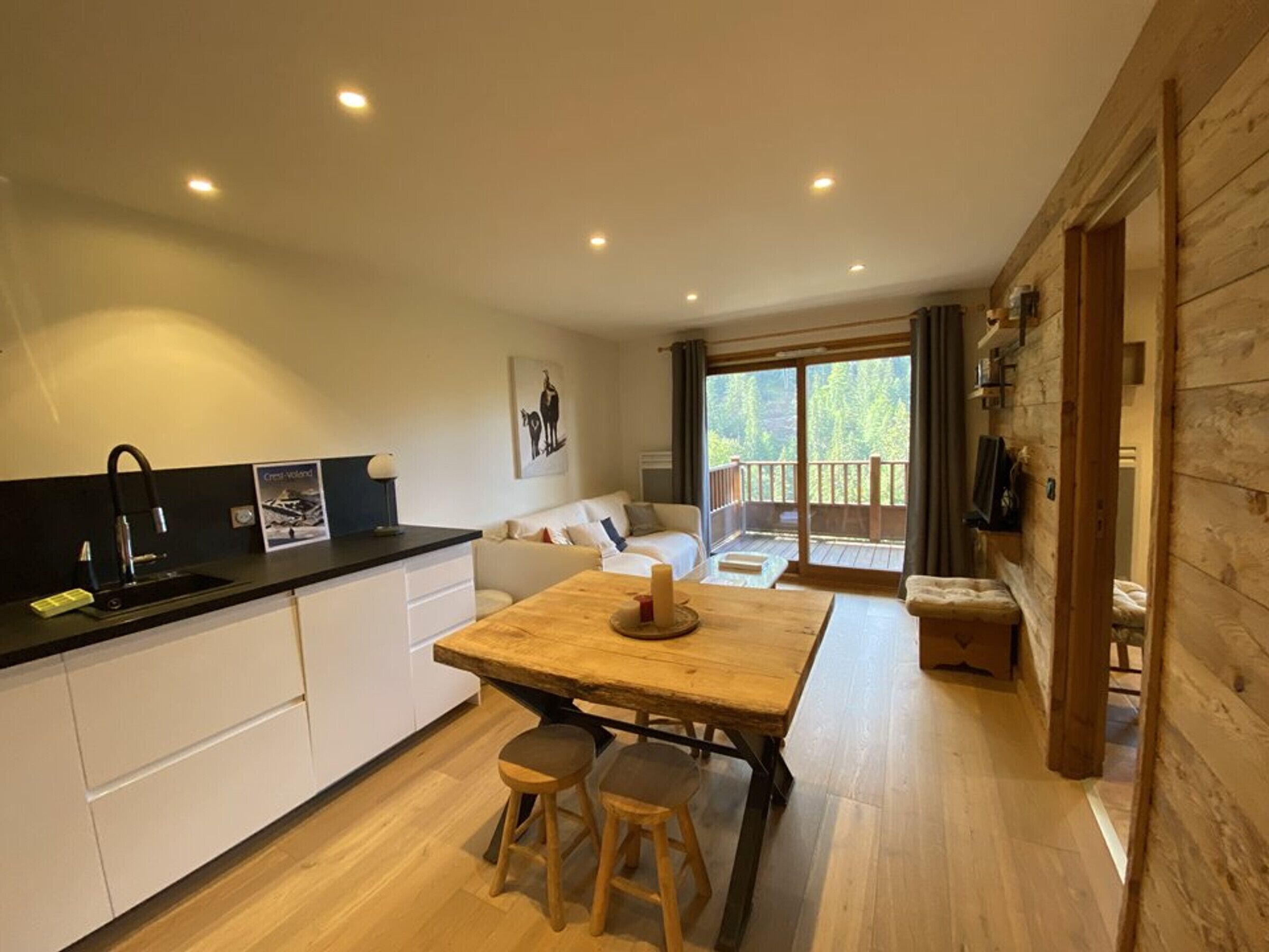 A modern kitchen with a wooden table and stools, a sofa, and a large window with a view of trees.