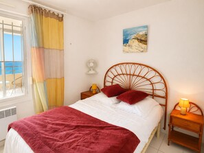 Ein Schlafzimmer mit einem Bett, einem Nachttisch, einem Wandventilator, einem Fenster mit Vorhängen und einem Bild.