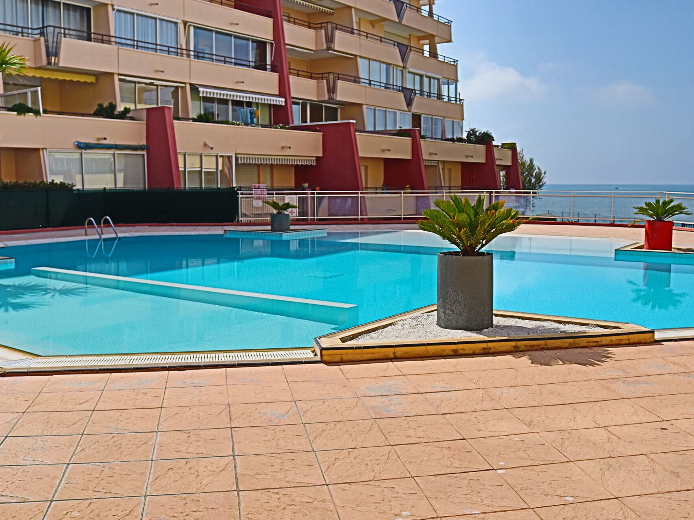 A swimming pool with a tiled deck, surrounded by a building with balconies and a view of the sea.